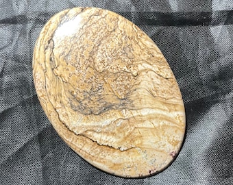 May include: An oval-shaped polished stone cabochon featuring a natural pattern of swirling tan, beige, and cream colours with dark brown linear inclusions and speckled textures throughout the surface, creating a landscape-like appearance.