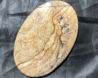 May include: An oval-shaped polished stone featuring a tan and beige base colour with intricate, flowing dark brown and black dendritic patterns that resemble a landscape or natural scenery across the smooth, glossy surface of the mineral.