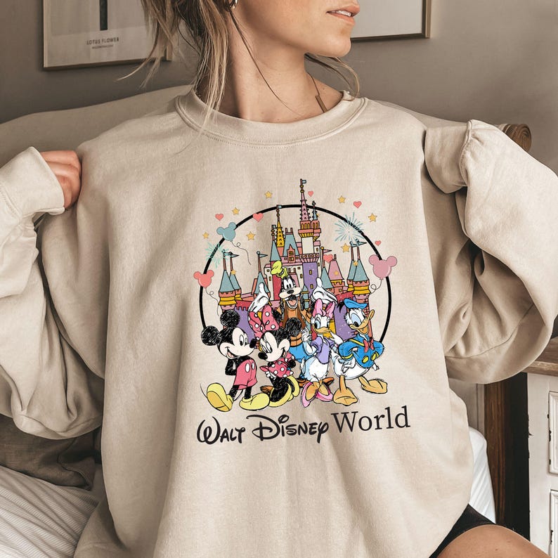 May include: Beige sweatshirt featuring a colorful graphic of Disney characters, including Mickey Mouse, Minnie Mouse, Donald Duck, and Daisy Duck, in front of a castle. The words "Walt Disney World" are printed below the graphic in a black script.