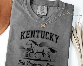May include: A gray Comfort Colors t-shirt featuring a black graphic of a horse, barrel, and the text "Kentucky The Bluegrass State EST. 1792." A gold necklace with a pendant rests on the shirt. A brown hat is visible in the upper left corner.