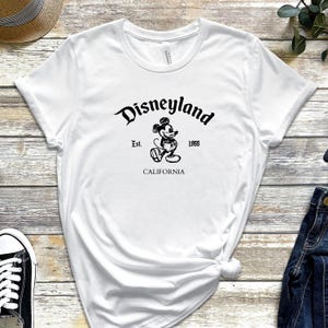 May include: White t-shirt featuring the Disneyland logo in black script with a Mickey Mouse graphic. The text includes "Est. 1955" and "CALIFORNIA". The shirt is made of a soft material.