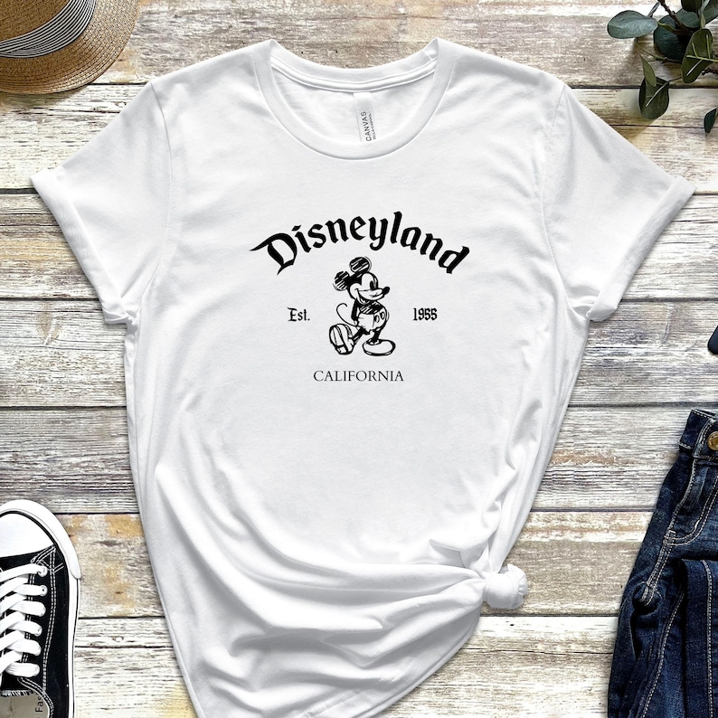 May include: White t-shirt featuring the Disneyland logo in black script with a Mickey Mouse graphic. The text includes "Est. 1955" and "CALIFORNIA". The shirt is made of a soft material.