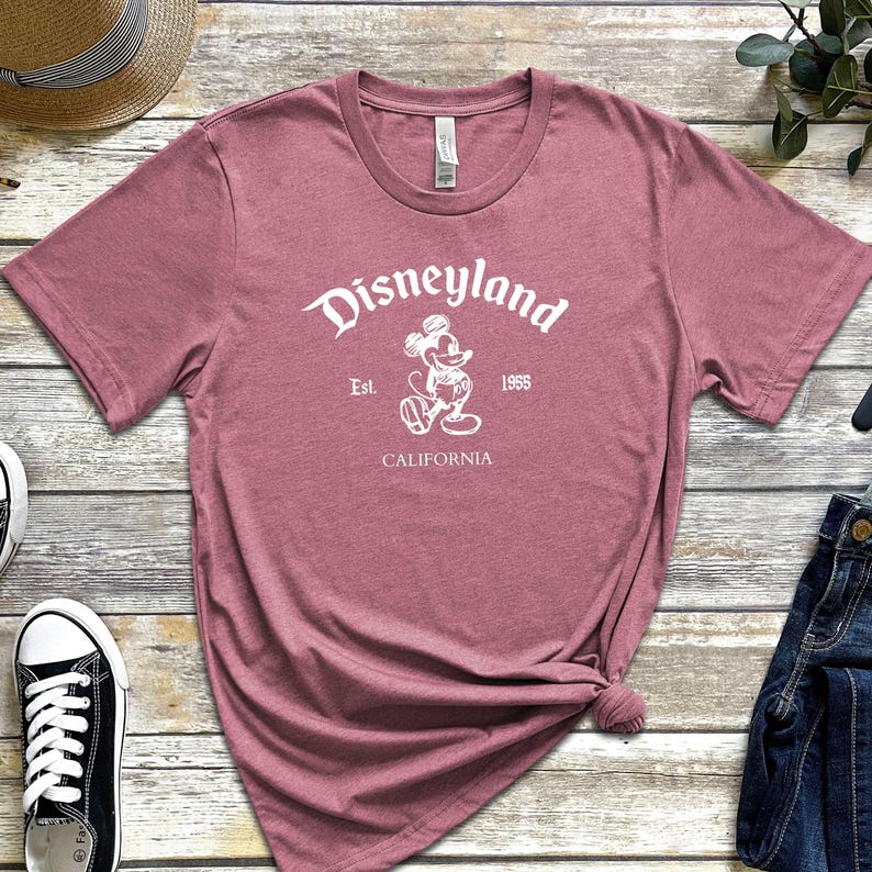May include: A dusty rose t-shirt with the Disneyland logo in white. The logo includes the word "Disneyland", the image of Mickey Mouse, the year "1955", and the word "California".