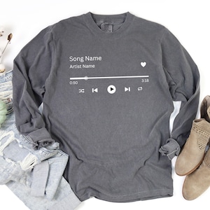 May include: A gray long-sleeve shirt featuring a white music player graphic. The graphic includes the text "Song Name" and "Artist Name", along with play, skip, and volume control icons. The shirt is displayed on a white surface.