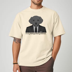 May include: Cream-colored t-shirt featuring a cartoon illustration of a dog wearing a suit and tie. The text "A very distinguished gentleman" is printed below the illustration. The shirt has short sleeves and a relaxed fit.