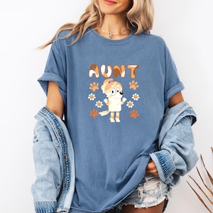 May include: A blue t-shirt with the word "AUNT" in brown and orange letters. The shirt features a cartoon dog character with daisies and paw prints. The shirt is made of a soft material.