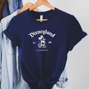 May include: Navy blue t-shirt with the word "Disneyland" in a script font, along with an image of Mickey Mouse. The text "Est. 1955" and "CALIFORNIA" are also printed on the shirt. The shirt is hanging on a wooden hanger.