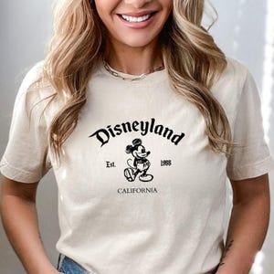 May include: Beige t-shirt featuring the word "Disneyland" in a stylized font, with "Est. 1955" and "CALIFORNIA" below. A black and white Mickey Mouse graphic is centered below the word "Disneyland".