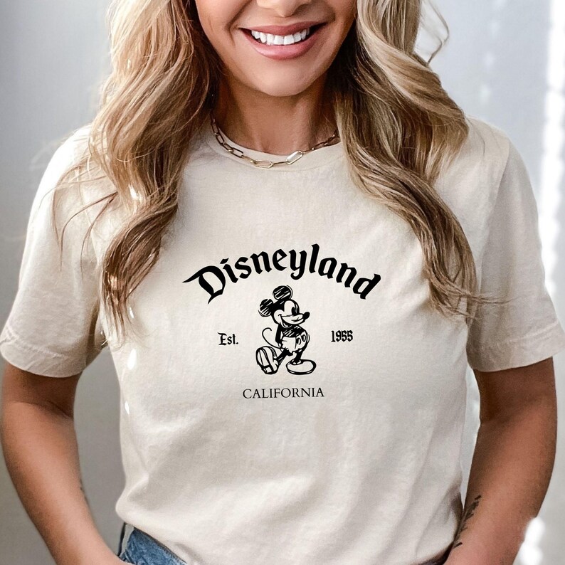 May include: Beige t-shirt featuring the word "Disneyland" in a stylized font, with "Est. 1955" and "CALIFORNIA" below. A black and white Mickey Mouse graphic is centered below the word "Disneyland".