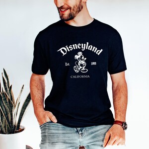 May include: A navy blue t-shirt featuring the Disneyland logo in white. The design includes the word "Disneyland" in a stylized font, the image of Mickey Mouse, and the text "Est. 1955 CALIFORNIA".