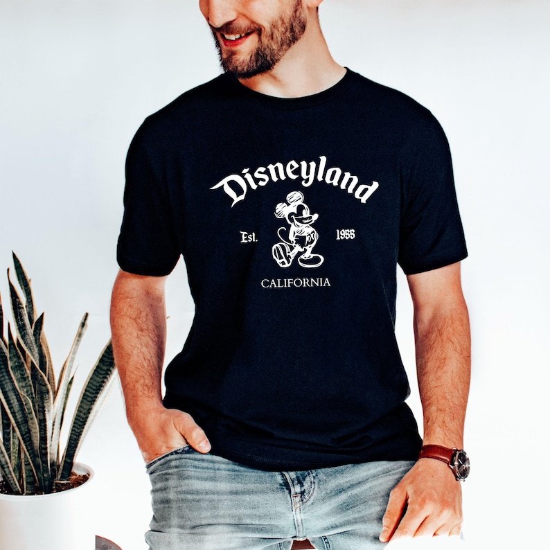 May include: A navy blue t-shirt featuring the Disneyland logo in white. The design includes the word "Disneyland" in a stylized font, the image of Mickey Mouse, and the text "Est. 1955 CALIFORNIA".