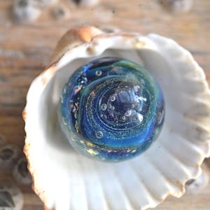 May include: A blue and green glass marble with gold flecks and air bubbles inside, resting in a white seashell.