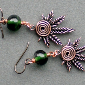 Könnte beinhalten: Ein Paar Ohrringe mit grünen Glasperlen und Designs aus Kupferdraht. Die Designs haben die Form einer Sonne mit einer Spirale in der Mitte.