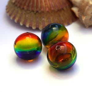 Könnte beinhalten: Drei kugelförmige Glasperlen mit einem lebendigen Regenbogenverlauf in Rot, Orange, Gelb, Grün, Blau und Violett. Jede Perle hat eine glatte, polierte Oberfläche und ein kleines Loch, das mittig zum Auffädeln durch die Perle gebohrt ist.