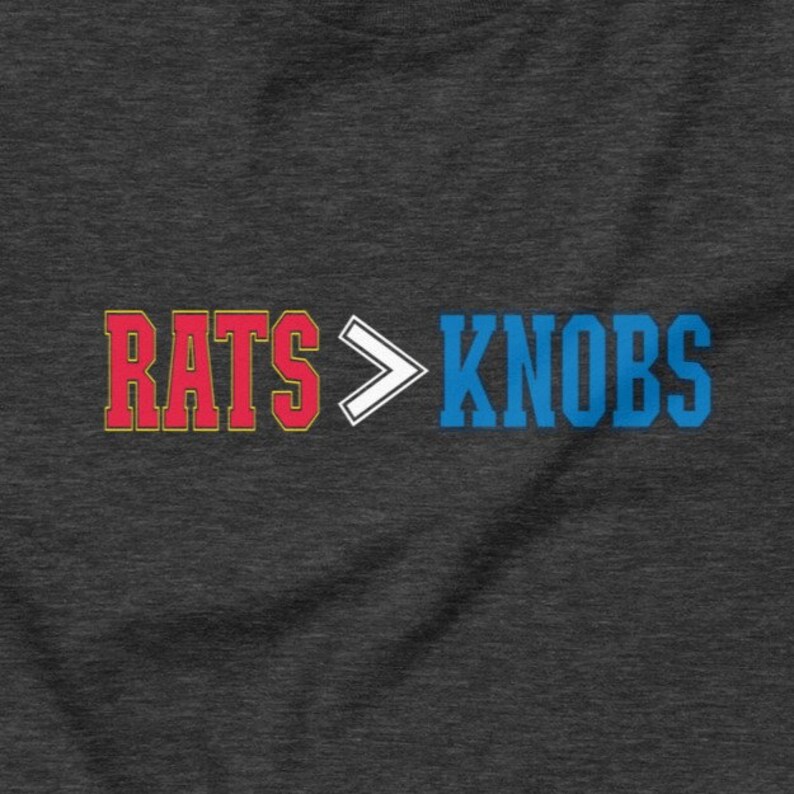 May include: Dark gray t-shirt with the text "RATS > KNOBS" in red and blue block letters. The word "RATS" is outlined in red and yellow, while "KNOBS" is in blue. A white arrow separates the words.