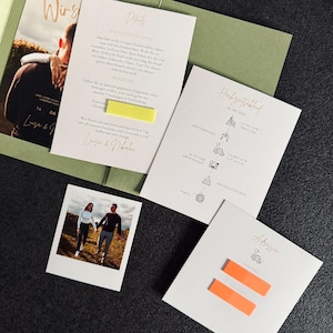 Könnte beinhalten: Hochzeitspapeterie-Set mit Einladungskarten und einem Foto. Die Karten sind weiß mit goldenem Text und orangefarbenen Haftnotizen. Eine Karte zeigt ein Foto eines Paares. Die Papeterie liegt auf einer dunklen Oberfläche.