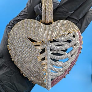 May include: A large, heart-shaped pendant with a gold bail and a detailed design. The pendant is encrusted with numerous clear and pink gemstones, with a ribcage design on one side. The pendant is held against a blue background.