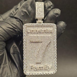 May include: A rectangular pendant with a large number 7 in the center, encrusted with small, sparkling stones. The pendant has a border of similar stones and the word "SEVEN" at the bottom. The top has a rectangular bail.