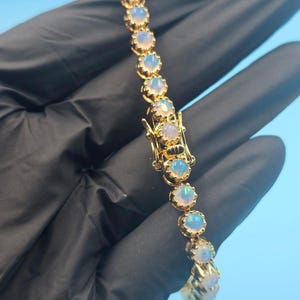 Op de afbeelding: Een gouden armband met een reeks ronde, iriserende opalen. Elke opaal is gezet in een gouden bezel en de armband is vastgezet met een gouden sluiting. De opalen vertonen een reeks kleuren, waaronder blauw, groen en geel.
