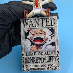 May include: A pendant featuring a cartoon character with a long red tongue, atop a "WANTED" poster. The poster has an image of a smiling character and the text "DEAD OR ALIVE? MONKEY D LUFFY". The pendant is embellished with rhinestones.