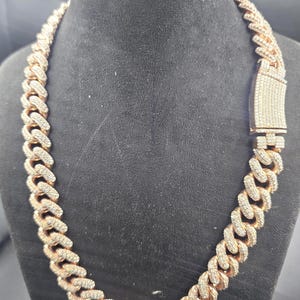 May include: A rose gold chain necklace with a cuban link design. The necklace is encrusted with small, clear gemstones. The clasp is rectangular and also covered in gemstones. The necklace is displayed on a black mannequin.