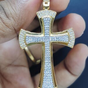 May include: Gold-toned pendant in the shape of a cross, encrusted with small, sparkling stones. The cross has a detailed design with a central section and edges lined with the stones. The pendant is held by a hand.