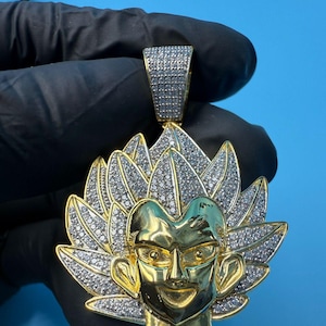 May include: A gold-coloured pendant featuring a stylised face with spiky hair, adorned with numerous small, clear gemstones. The pendant has a bail at the top for a chain. The design is set against a blue background.