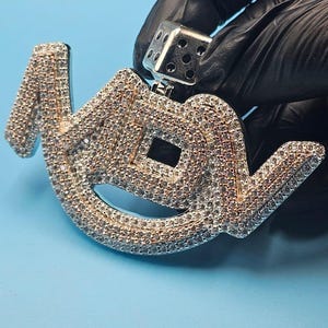 May include: A silver pendant spelling "MDV", studded with small, sparkling stones. The pendant is held by a silver die-shaped bail. The background is a solid light blue.