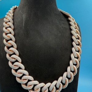 May include: A rose gold chain necklace with large, interlocking links, encrusted with small, sparkling stones. The necklace is displayed on a black mannequin against a blue background, showcasing its bold design and luxurious appearance.