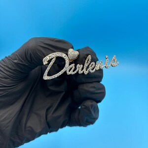 May include: A gold-colored pendant with the name "Darlenis" spelled out in a cursive font, encrusted with small, clear gemstones. A small heart shape is placed above the "D". The pendant is held against a blue background.