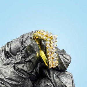 May include: A gold ring featuring a V-shaped chevron design adorned with two rows of clear, round-cut stones. The band is crafted from a polished yellow metal, and the stones are set in a prong setting, creating a sparkling, layered appearance.