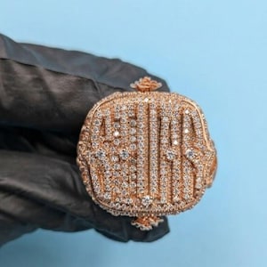 May include: A rose gold ring featuring a square-shaped face covered in numerous small, clear, sparkling stones. The design includes vertical rows of stones and a decorative floral-like element at the top and bottom of the ring's face.