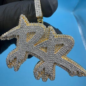 May include: A gold-toned pendant with a stylized double-letter design, encrusted with numerous small, sparkling white stones. The pendant has a bail at the top for a chain. The background is a solid light blue.