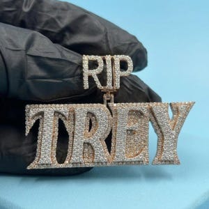 May include: A gold-toned pendant with the letters "RIP TREY" encrusted with small, sparkling stones. The pendant is held against a dark background, showcasing the intricate details and design of the jewelry.