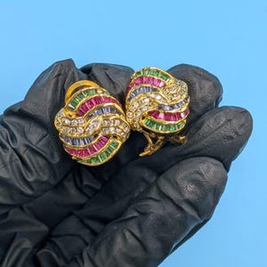 Könnte beinhalten: Ein Paar goldfarbene Clip-Ohrringe. Jeder Ohrring hat ein wirbelndes Design mit abwechselnden Reihen aus roten, grünen, blauen und weißen Edelsteinen. Die Ohrringe werden von einer behandschuhten Hand vor blauem Hintergrund gehalten.