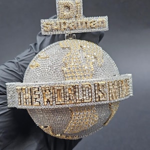 May include: A diamond-encrusted pendant featuring a globe design with gold continents and the phrase "THE WORLD IS MINE." The pendant also includes a cross-shaped top with the inscription "DI SUPREME."