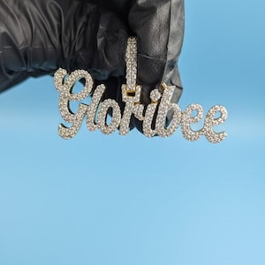 May include: A gold-colored pendant spelling "Gloribee" in a cursive font, encrusted with small, clear gemstones. The pendant is held against a light blue background.