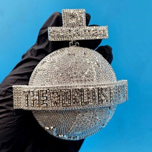 以下が含まれることがあります： 「THE WORLD IS YOURS」の文字が太字で刻まれた、ダイヤモンドをちりばめたペンダント。ペンダントは地球儀のデザインで、十字型のトップがあり、すべて青い背景に設定されています。