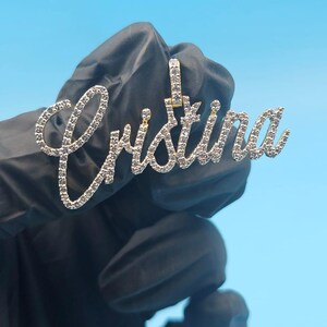 May include: A gold pendant spelling "Cristina" in a cursive font, set with small, clear gemstones. The pendant is held against a blue background.