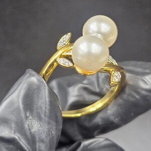 Può includere: Un anello d'oro con due grandi perle luminose. La fascia dell'anello è decorata con delicati motivi a forma di foglia, impreziositi da piccoli diamanti scintillanti. L'anello è posto su uno sfondo scuro.