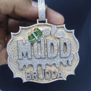 May include: A gold and silver pendant with the word "MUDD" and "BRUDDA" in silver, encrusted with small, sparkling stones. The pendant features a green and gold money design and a decorative border.