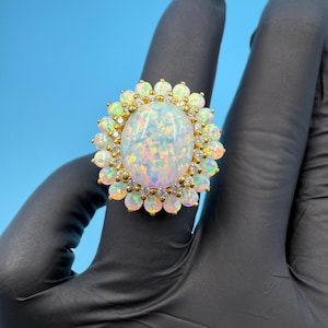 May include: A gold ring featuring a large, oval opal gemstone surrounded by smaller opals and clear crystals. The ring has a floral design with a gold band and is set against a blue background.