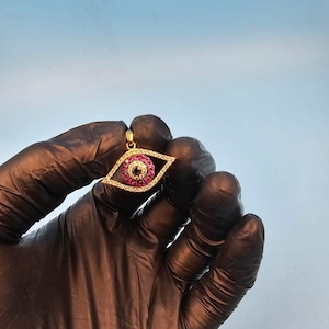 May include: A gold-colored pendant in the shape of an eye, encrusted with small red and clear gemstones. The center of the eye features a black gemstone. The pendant is held against a light blue background.