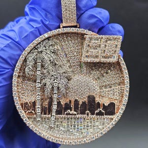 May include: A round, rose gold pendant encrusted with sparkling gemstones. The pendant features a detailed cityscape with palm trees, a dome, and a flag. The outer edge is lined with smaller gemstones.