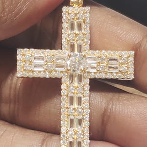 May include: A gold-coloured cross pendant, adorned with many small, sparkling diamonds. The cross has a central, larger diamond and baguette-cut diamonds. The pendant is held by a hand.
