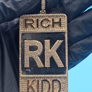 以下が含まれることがあります： 「RICH」と「KIDD」の文字が浮き彫りになったゴールドトーンのペンダント。中央には大きな文字で「RK」が配置されています。ペンダントは長方形で、中央は黒色で、小さな輝く石がちりばめられています。