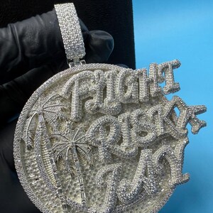 May include: A silver-colored pendant with the words "Flight Risk Day" in a stylized font. The pendant features a round design with palm trees and is encrusted with small, sparkling stones. The bail is also covered in stones.