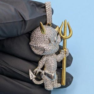 May include: A diamond-encrusted pendant in the shape of a cartoon devil holding a gold trident. The devil has gold horns and a silver diaper. The pendant is set against a light blue background.