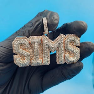 Puede incluir: Un colgante dorado que deletrea el nombre "SIMS" en letras de bloque. Las letras están incrustadas con pequeñas piedras brillantes, creando un efecto deslumbrante. El colgante se sostiene sobre un fondo azul brillante.
