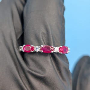 May include: A silver ring with a row of oval-cut, deep red rubies and smaller, clear gemstones. The ring is set against a black background with a light blue backdrop.